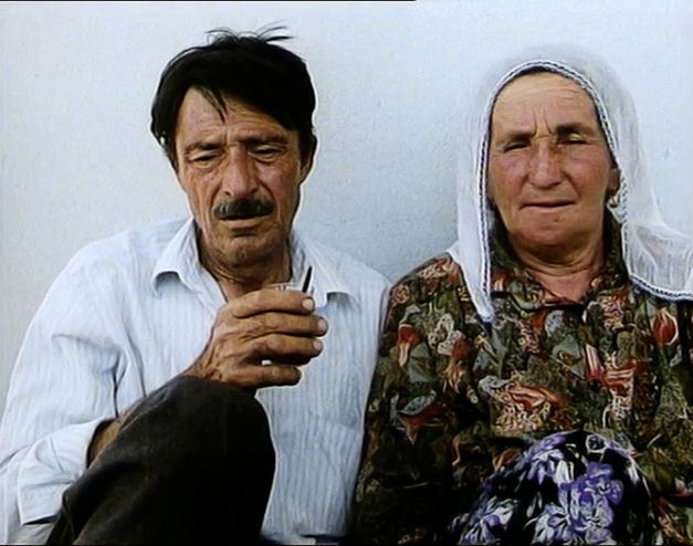 Still from the film „Mein Vater, der Gastarbeiter“ by Yüksel Yavuz. On the left side, a man with a moustache sits against a white wall and holds a glass in his hand. On the right side sits a woman with a cloth on her head.