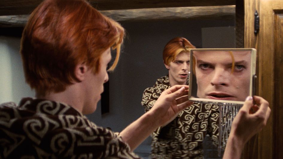 Film still from THE MAN WHO FELL TO EARTH: David Bowie looks at himself in both a large and a small mirror at the same time, so that he appears three times.