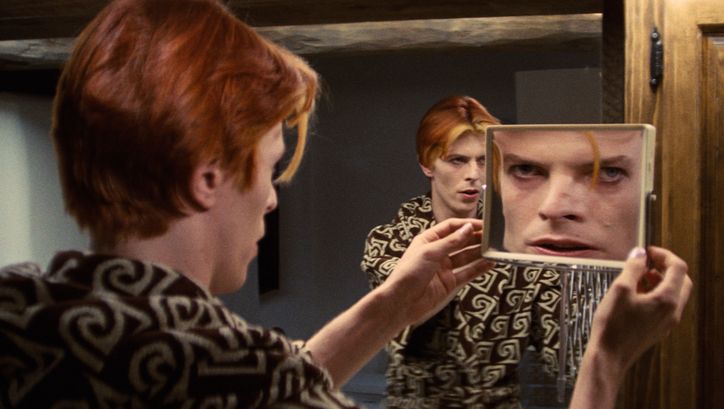 Film still from THE MAN WHO FELL TO EARTH: David Bowie looks at himself in both a large and a small mirror at the same time, so that he appears three times.