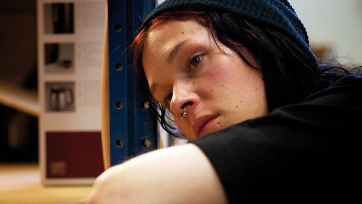 Filmstill aus SOMETHING MUST BREAK: Eine junge Person lehnt sich auf einen Tisch und blickt nachdenklich in die Ferne.