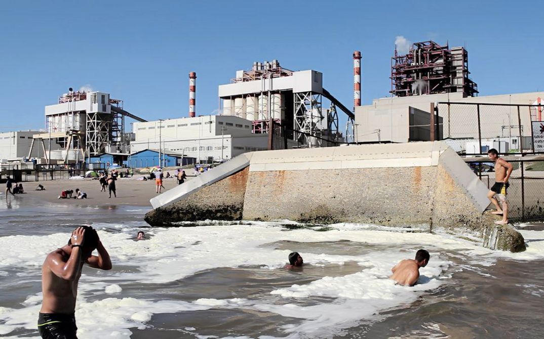 Filmstil aus "Oasis" von Tamara Uribe und Felipe Morgado. Zu sehen ist ein Strand direkt neben einem Industriegelände. Menschen schwimmen im Wasser und sitzen am Strand. 