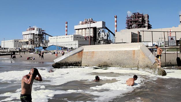 Film stil from "Oasis" by Tamara Uribe and Felipe Morgado. It shows a beach right next to an industrial site. People are swimming in the water and sitting on the beach. 
