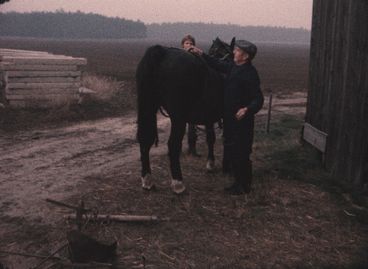 Ein Mann und eine Frau stehen an zwei Seiten eines schwarzen Pferdes, das von der Kamera wegschaut. Sie stehen an einer Scheune, und im Hintergrund ist ein Feld zu sehen. Das Bild hat einen ausgeprägten rötlichen Farbton.