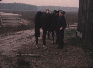 A man and a woman stand on two sides of a black horse, which is facing away from the camera. They are beside the wall of a barn, and we see a field in the background. The image has a pronounced reddish tint.