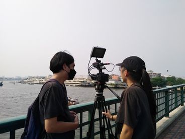 A behind-the-scenes shot of two people. One is filming and the other is standing next to them, wearing a face mask. There is a large body of water in front of them, and they seem to be standing on a bridge.
