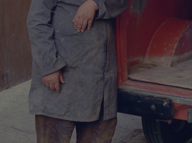 Film still from Hicham Gardaf’s film “In Praise of Slowness”. A person from the chest-down, wearing a gray, stained shirt, leaning on what seems to be the back of a red truck. 