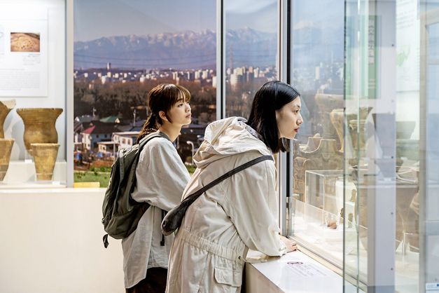 Zwei junge Frauen in weißer Kleidung sind im Profil zu sehen, sie stehen in einem Museum und schauen sich Ausstellungsstücke in einer Vitrine an. Im Hintergrund ist durch ein großes Fenster ein Panorama mit einer Stadt und Bergen zu sehen..