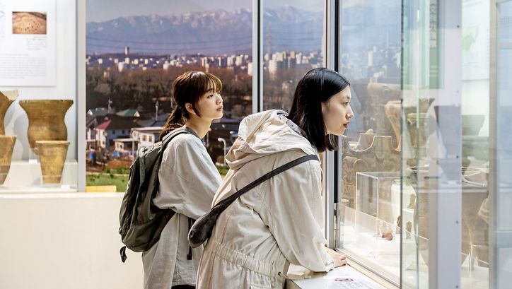 Zwei junge Frauen in weißer Kleidung sind im Profil zu sehen, sie stehen in einem Museum und schauen sich Ausstellungsstücke in einer Vitrine an. Im Hintergrund ist durch ein großes Fenster ein Panorama mit einer Stadt und Bergen zu sehen..