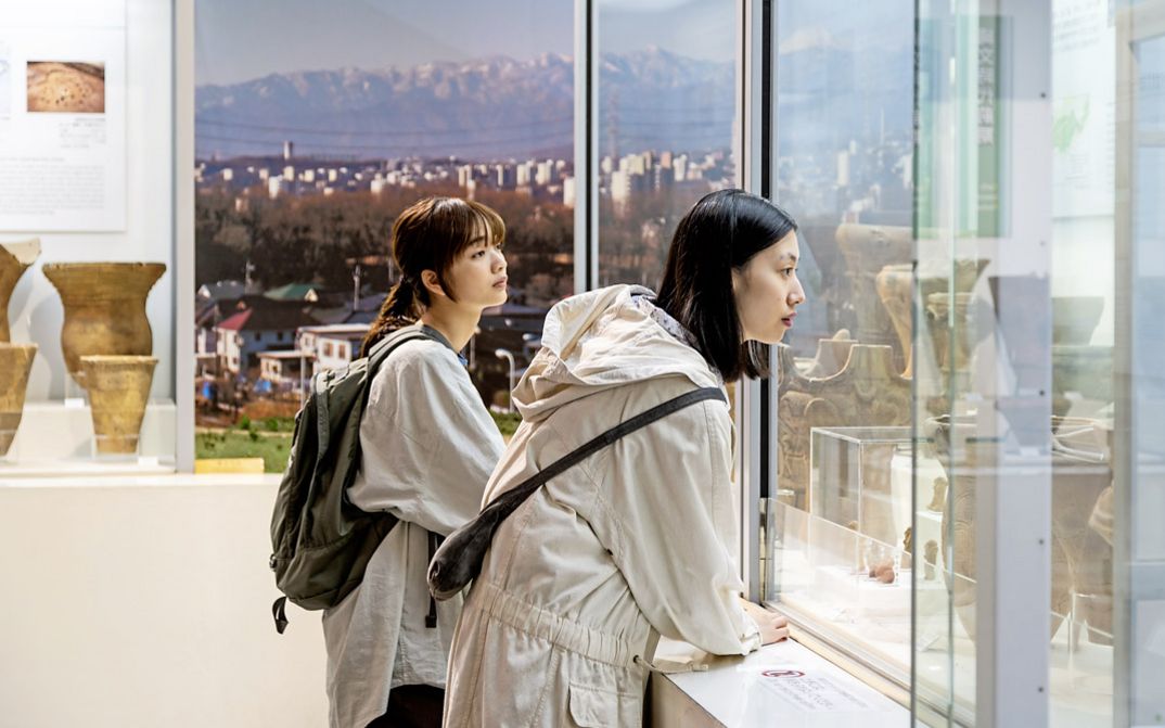 Zwei junge Frauen in weißer Kleidung sind im Profil zu sehen, sie stehen in einem Museum und schauen sich Ausstellungsstücke in einer Vitrine an. Im Hintergrund ist durch ein großes Fenster ein Panorama mit einer Stadt und Bergen zu sehen..