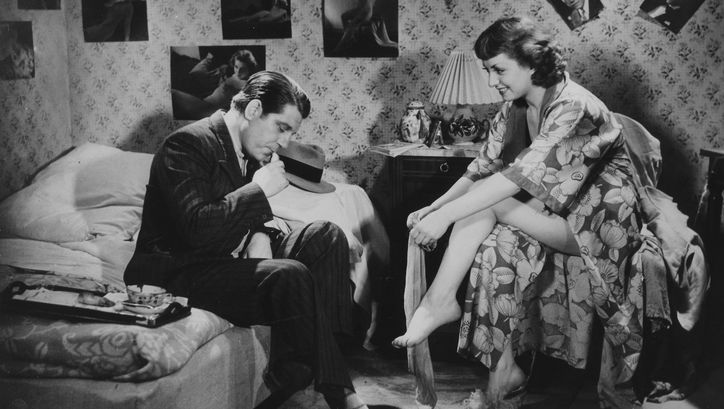 Film still from LA BELLE EQUIPE: In a bedroom, the walls are covered with posters of celebrities. A man in a suit is sitting on the bed. Opposite him, on a chair, sits a woman in a bathrobe putting on stockings.