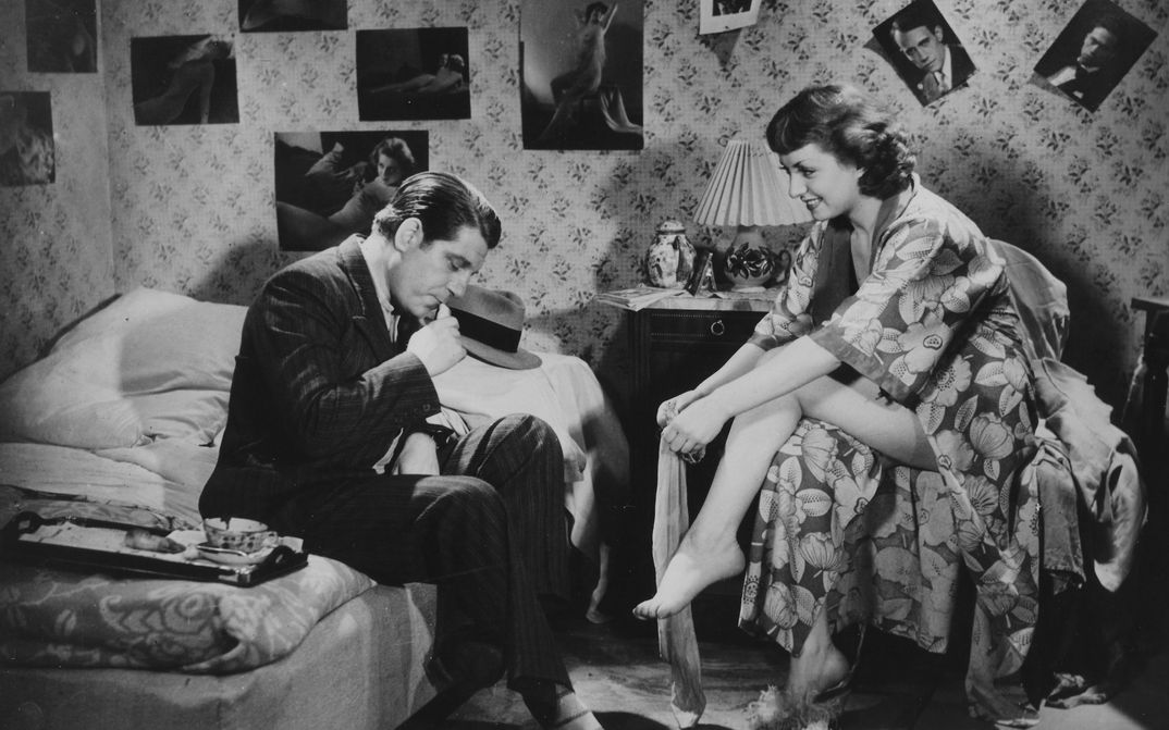 Film still from LA BELLE EQUIPE: In a bedroom, the walls are covered with posters of celebrities. A man in a suit is sitting on the bed. Opposite him, on a chair, sits a woman in a bathrobe putting on stockings.