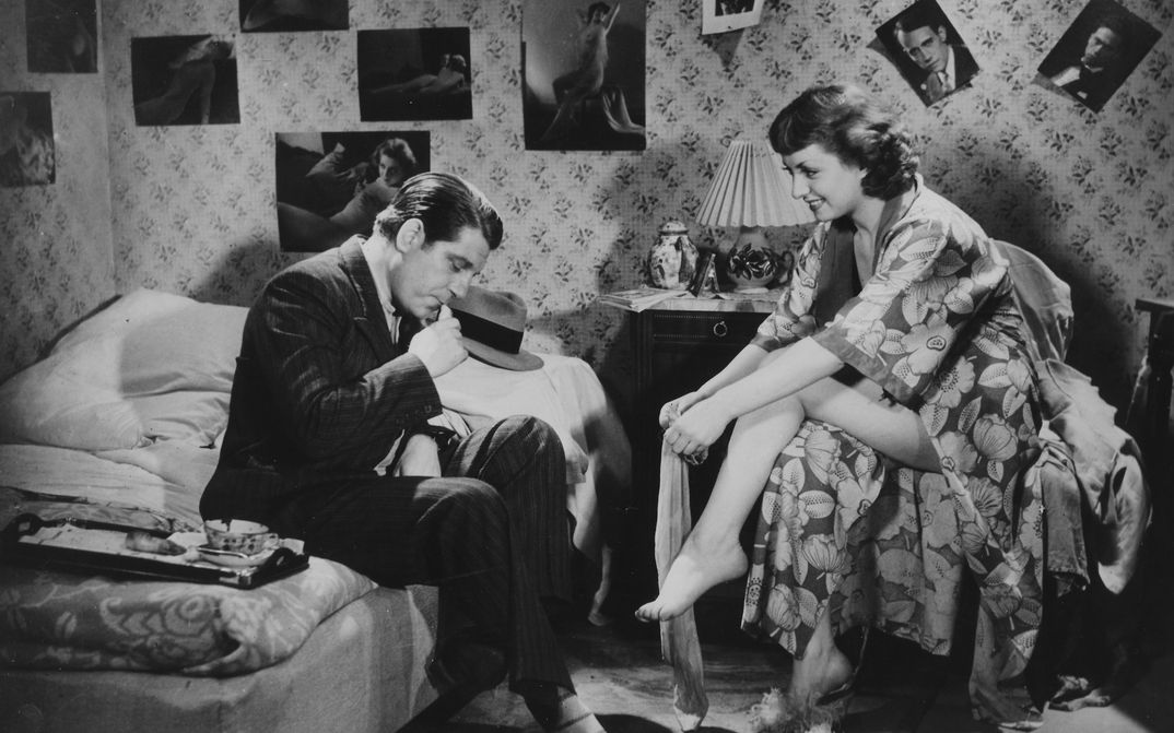 Filmstill aus LA BELLE EQUIPE: In einem Schlafzimmer sind die Wände voller Poster von Stars. Ein Mann im Anzug sitzt auf dem Bett. Ihm gegenüber auf einem Stuhl sitzt eine Frau im Bademantel, die sich Strümpfe anzieht.