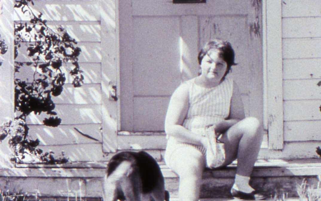 Film still from LITTLE GIRL. A girl sits with her dog on the steps in front of a house.