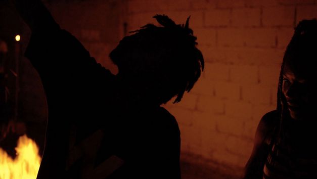 Still from the film "The Wake" by The Living and the Dead Ensemble. The silhouette of a person in the dark in front of a brick wall.