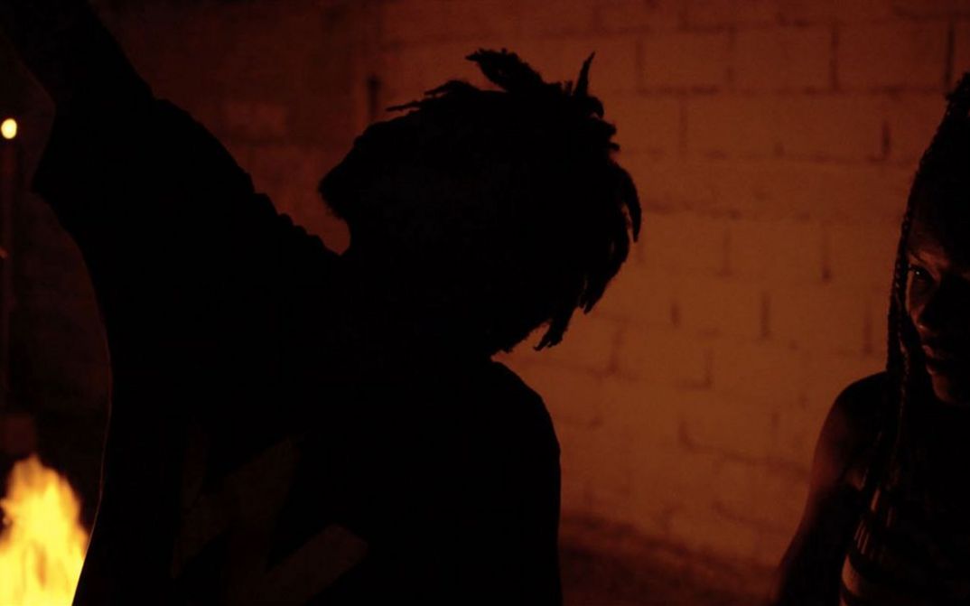 Filmstill aus dem Film "The Wake" von The Living and the Dead Ensemble. Man sieht die Silhouette einer Person im Dunkeln vor einer Ziegelwand.