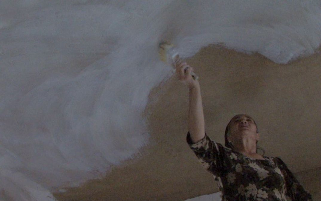 Film still from BARZAKH: A woman paints a ceiling with white paint.