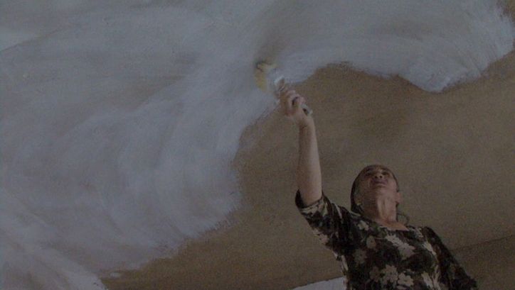 Film still from BARZAKH: A woman paints a ceiling with white paint.