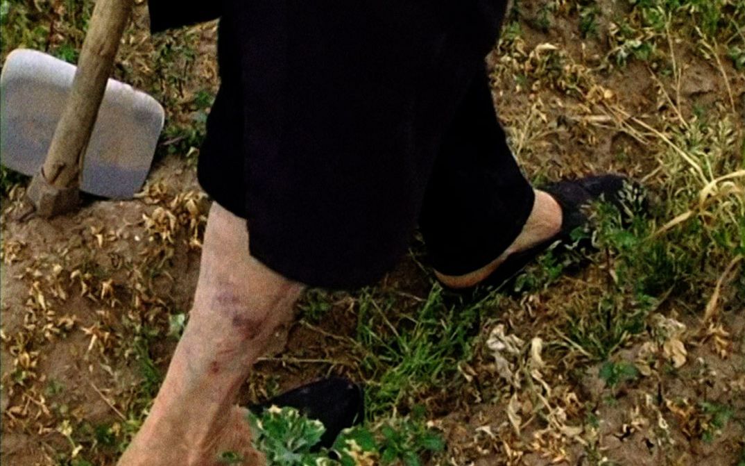 Filmstill aus „Terra que marca (Striking Land)“ von Raul Domingues. Großaufnahme der Beine einer Bäuerin, die über das Feld geht. Links oben ist eine Hacke zu sehen.