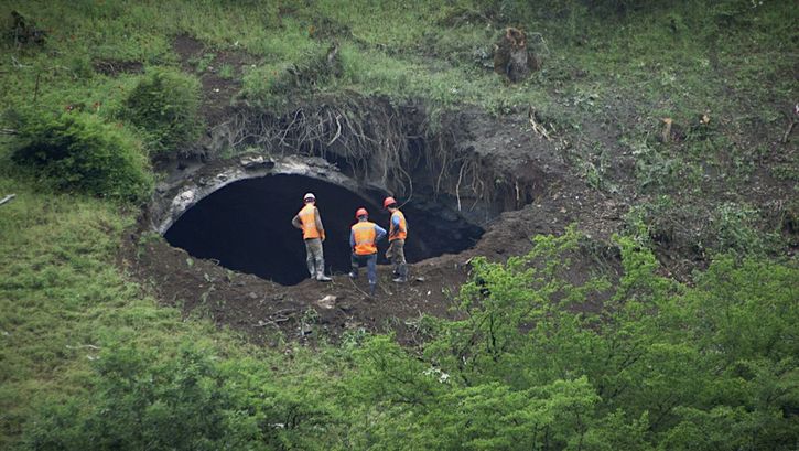 Filmstill aus A TUNNEL. Drei Männer in Arbeitskleidung schauen in eine Tunnelöffnung inmitten einer grünen Landschaft.