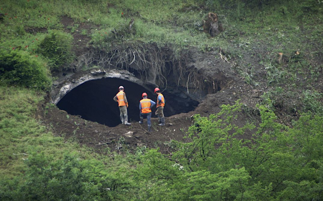 Filmstill aus A TUNNEL. Drei Männer in Arbeitskleidung schauen in eine Tunnelöffnung inmitten einer grünen Landschaft.