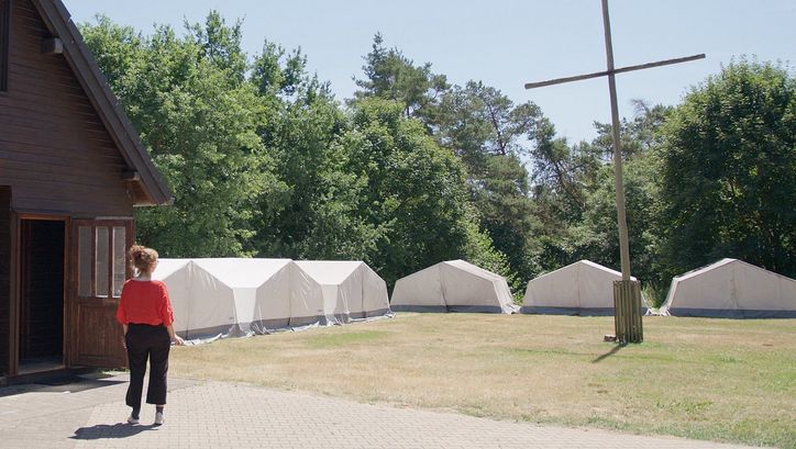 Film still from ZWISCHEN UNS GOTT: On the left side of the picture, you can see a woman from behind. She is looking at a meadow with a row of white tents and a large cross.