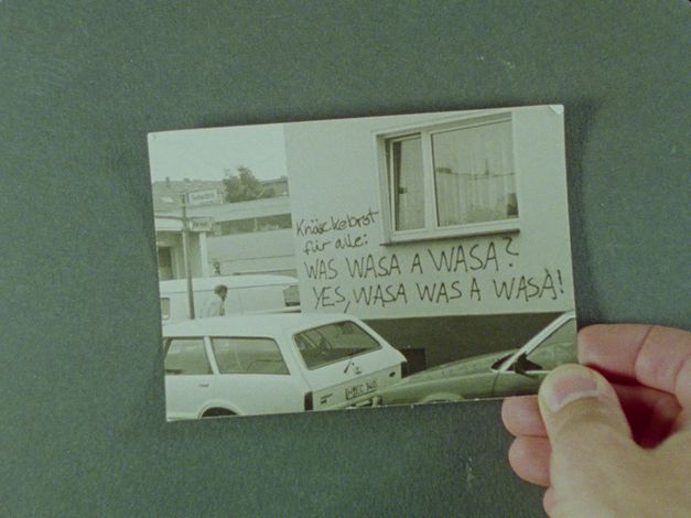 Film still from EIGENTLICH EIGENTLICH JANUAR: A hand holds a photograph of cars parked in front of a house inscribed with graffiti.
