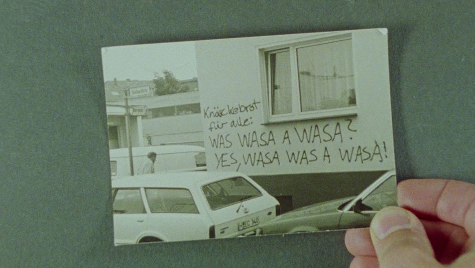 Film still from EIGENTLICH EIGENTLICH JANUAR: A hand holds a photograph of cars parked in front of a house inscribed with graffiti.