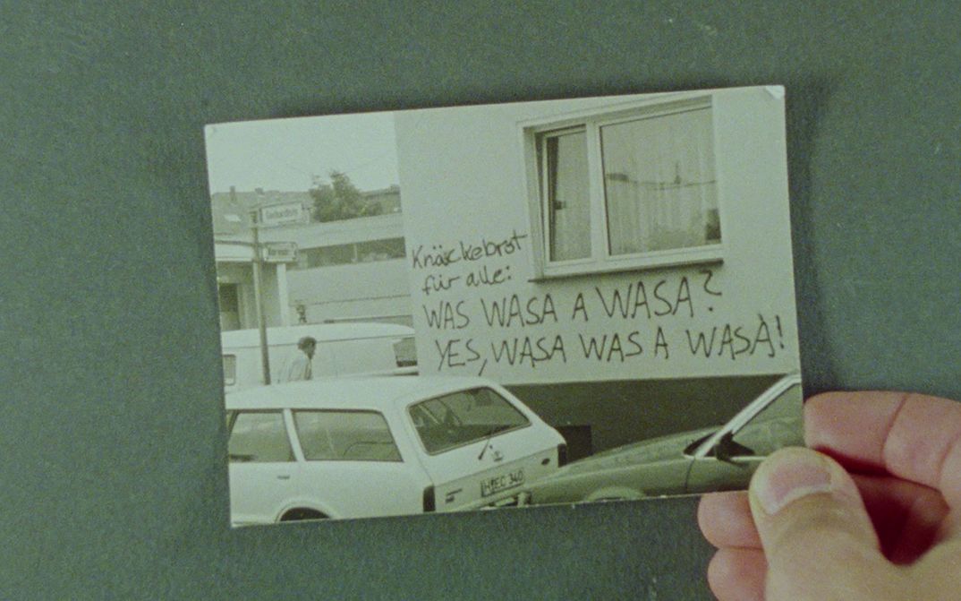 Filmstill aus EIGENTLICH EIGENTLICH JANUAR: Eine Hand hält eine Fotografie, auf der Autos vor einem mit Grafitti beschrifteten Haus stehen.