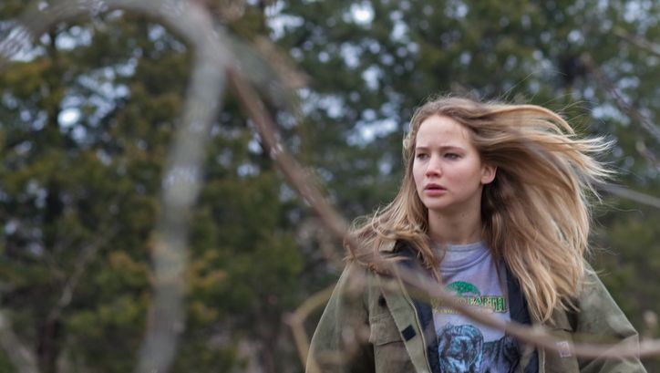 A young woman with long blonde hair and a determined expression stands among the trees; in the foreground of the picture, blurred branches can be seen.Film still from WINTER