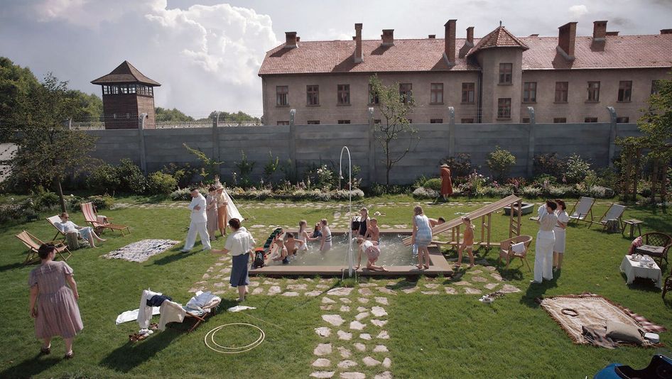 Film still from THE ZONE OF INTEREST: A garden with a small pool is filled with adults and children swimming and having fun. In the background, you can see a fence topped with barbed wire, and behind it, a watchtower from the Auschwitz concentration camp.