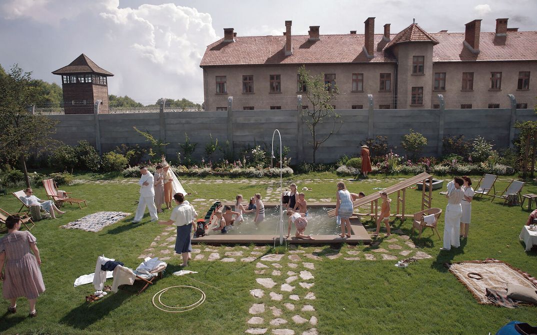 Filmstill aus THE ZONE OF INTEREST: Ein Garten mit einem kleinen Pool ist voller Erwachsener und Kinder, die baden und sich amüsieren. Im Hintergrund sieht man einen Zaun mit Stacheldraht, dahinter ein Wachturm des Konzentrationslagers Auschwitz.