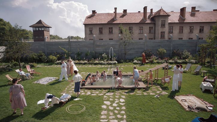 Filmstill aus THE ZONE OF INTEREST: Ein Garten mit einem kleinen Pool ist voller Erwachsener und Kinder, die baden und sich amüsieren. Im Hintergrund sieht man einen Zaun mit Stacheldraht, dahinter ein Wachturm des Konzentrationslagers Auschwitz.
