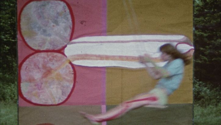 Filmstill aus JÜM JÜM: Eine junge Frau beim Schaukeln. Dahinter ein aufgehängtes Tuch mit buntem Muster.