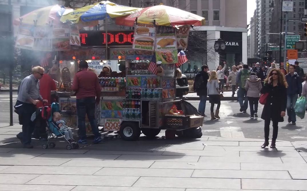 Filmstill aus POPEYE SEES 3D: Eine Straßenszene in New York. Man sieht einen Hot-Dog-Verkaufswagen und Menschen über eine Kreuzung gehen.