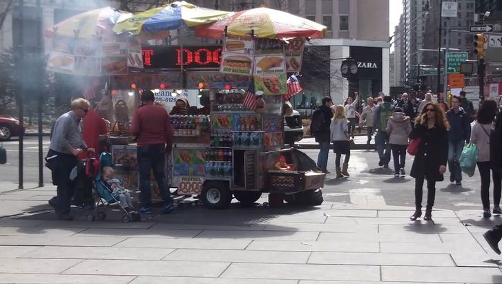 Filmstill aus POPEYE SEES 3D: Eine Straßenszene in New York. Man sieht einen Hot-Dog-Verkaufswagen und Menschen über eine Kreuzung gehen.