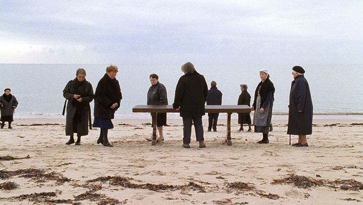 Film still from QUELQUE VEUVES DE NOIRMOUTIER. Some older women dressed in black are standing on a beach, in their midst is a table.