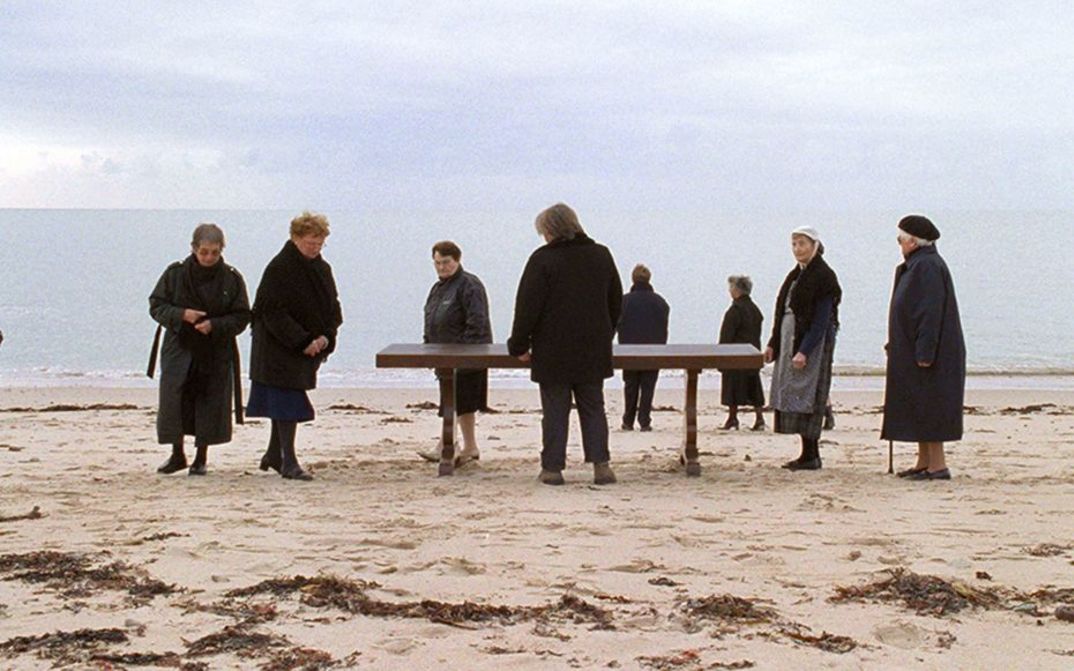 Film still from QUELQUE VEUVES DE NOIRMOUTIER. Some older women dressed in black are standing on a beach, in their midst is a table.