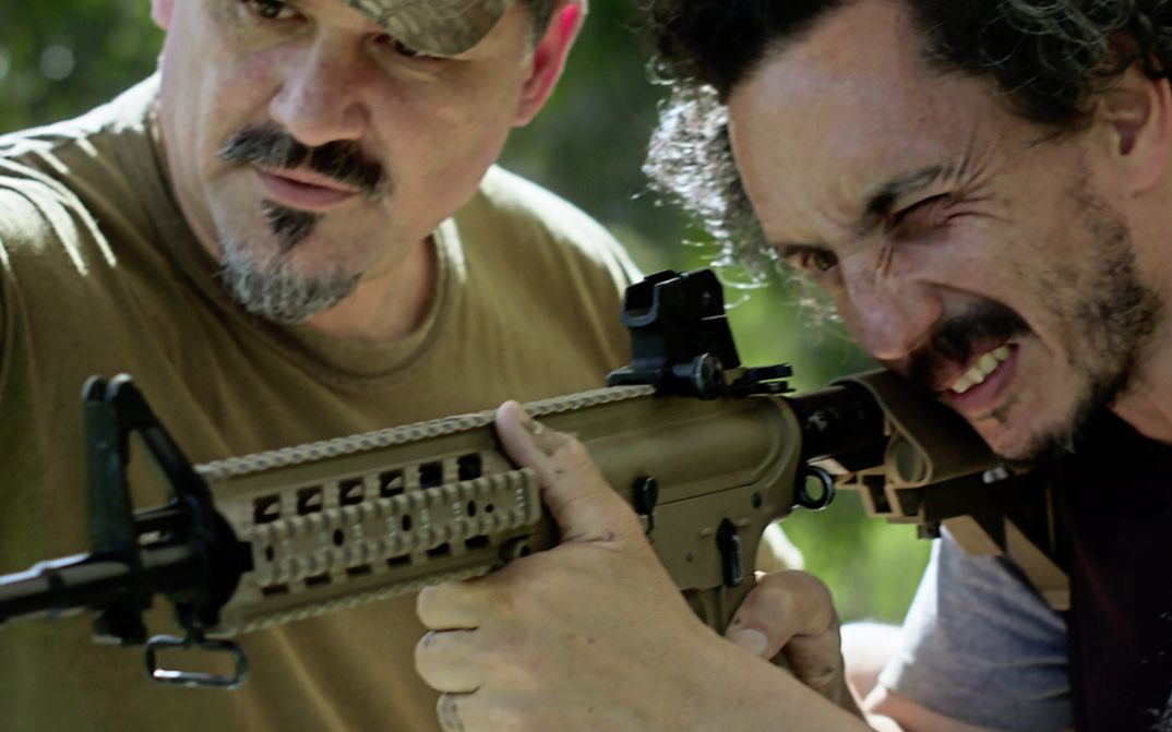 Still from the film "Camuflaje (Camouflage)" by Jonathan Perel. Two men in medium-close up look down the barrel of an automatic rifle. 