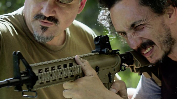 Still from the film "Camuflaje (Camouflage)" by Jonathan Perel. Two men in medium-close up look down the barrel of an automatic rifle. 