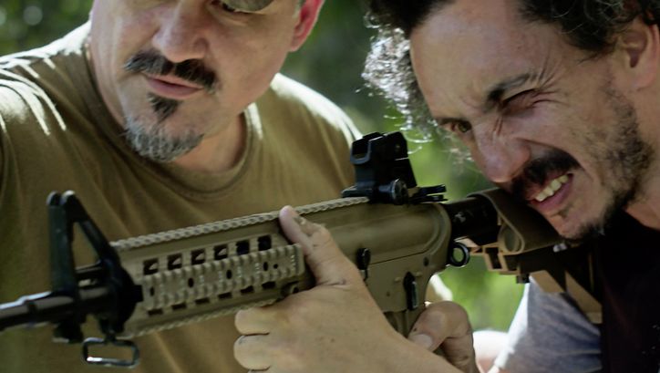 Still from the film "Camuflaje (Camouflage)" by Jonathan Perel. Two men in medium-close up look down the barrel of an automatic rifle. 