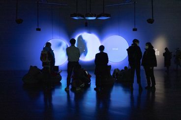 In a dark exhibition space, a few people are standing in front of three round screens, watching. Everything appears in a blue light.