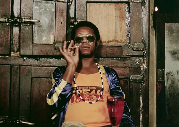 A man with sunglasses and a long black and white necklace holds a joint between his fingers. He stands with his back to an old wooden cupboard with many massive doors.