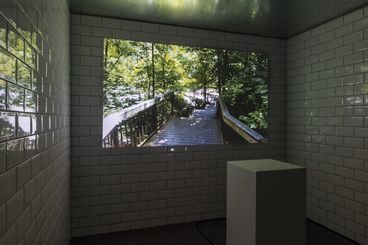 In a white tiled room a video is projected on the wall. In the center of the room is a white pedestal. 