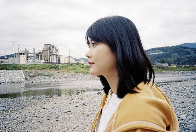 Still from the film "Ishi ga aru" by Tatsunari Ota. We can see the profile of a woman with black hair and a yellow jacket. There is a factory-like building in the background, and a stream of water surrounded by rocks in between the building and the woman.