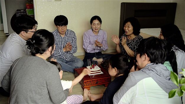 Still from the film "Hot in Day, Cold at Night" by Park Song-yeol. We see a group of people sitting and clapping on the ground around a birthday cake.