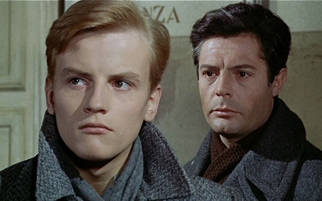 Film still from CRONACA FAMILIARE: Two men in winter coats are standing one behind the other, looking in the same direction.