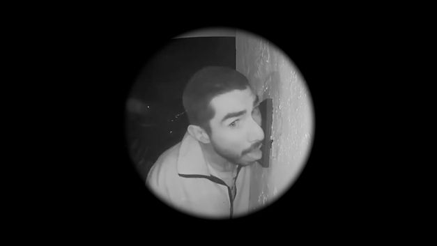 Film still from Graeme Arnfield’s „Home Invasion“. A black and white circular view of a person licking a doorbell on the wall on the right side.