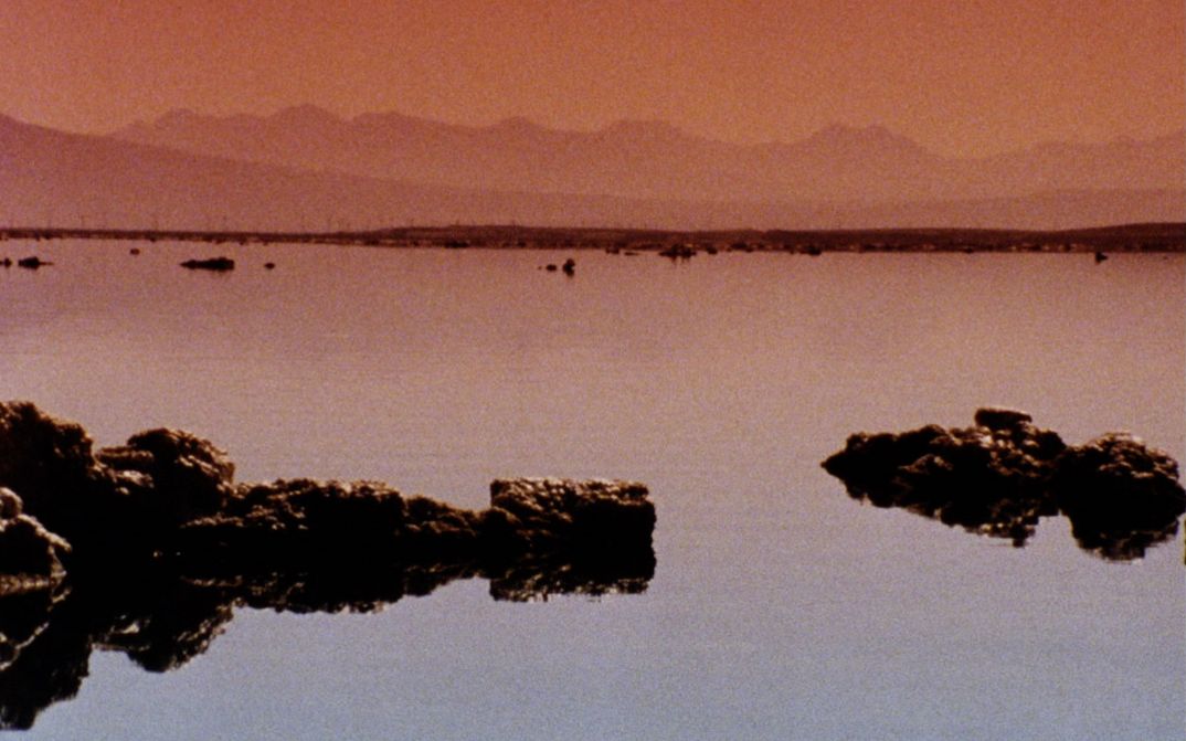 Filmstill aus dem Film "Instant Life" von Anja Dornieden, Juan David González Monroy und Andrew Kim. Zu sehen ist ein Gewässer mit Felsstrukturen, im Hintergrund Berge.