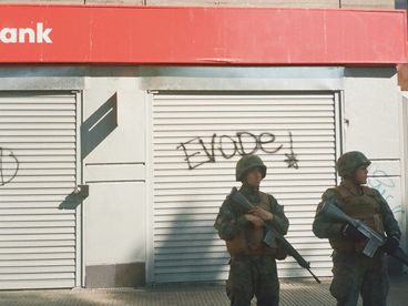 35mm colour photo of two men in military gear with guns standing in front of a shuttered bank, with the word “evade!” spray painted on façade.