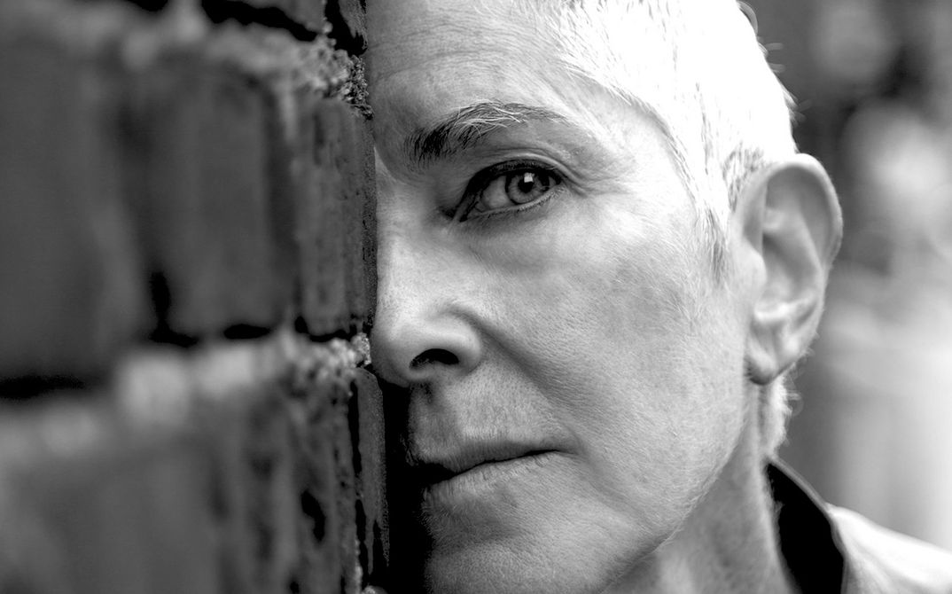 Porträtfoto von Beth B. Sie trägt kurze Haare und schaut direkt in die Kamera. Teile ihres Gesichts sind hinter einer Mauer versteckt.